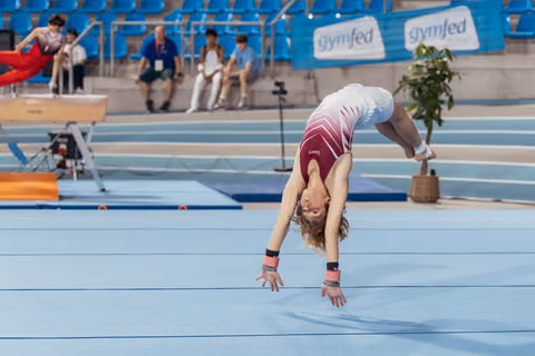 Young gymnast performs a dramatic back walkover on the blue floor, arched deeply with hair flowing, at a Gymfed competition.