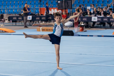Young male gymnast performs an arabesque on the floor exercise, arms outstretched, focused expression in a competition arena.