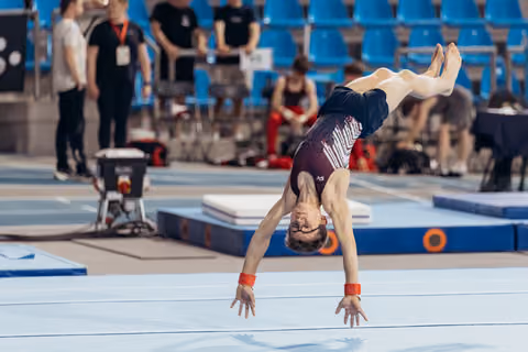 Young gymnast performing an inverted handstand on floor, wearing glasses and red wristbands, focused expression mid-routine.