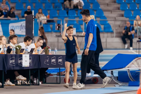 Smiling young gymnast waves to judges while walking with coach in blue at a gymfed gymnastics event.