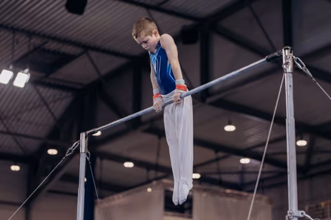Young male gymnast grips the high bar with intense focus, body suspended mid-routine in an indoor gymnasium.