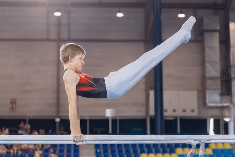 Young male gymnast holds a straddle L-sit on parallel bars, legs raised high, expression intense and focused, in a sports hall.