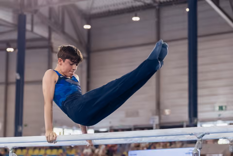 Young male gymnast performs a focused pike hold on parallel bars in a large indoor sports hall.