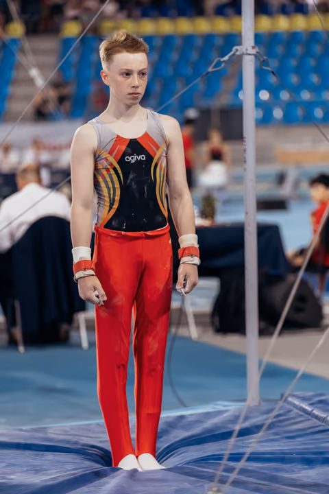 Young male gymnast stands composed and focused near high bar apparatus, wearing red pants and agiva leotard.