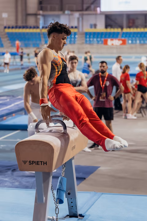Young male gymnast performs on pommel horse, focused expression, wearing orange-black leotard in busy arena.