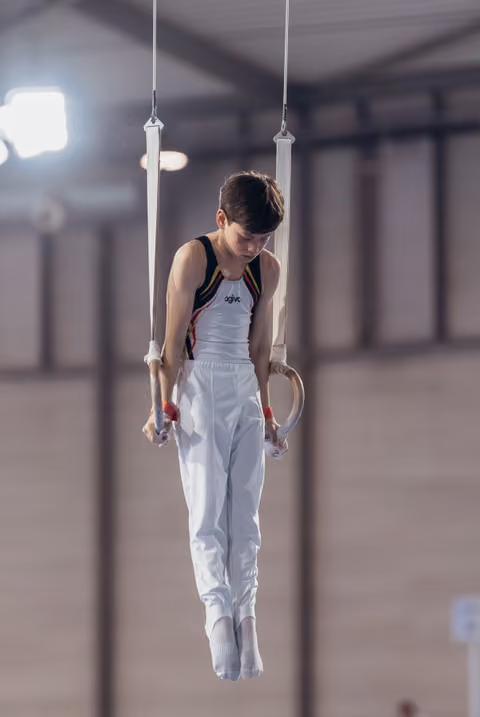 Young male gymnast hangs still on rings, head bowed in concentration, wearing white and black competition leotard indoors.