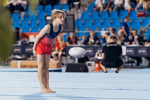 Young gymnast stands barefoot on floor mat, leaning forward with intense focus, preparing for her routine at a gymnastics competition.
