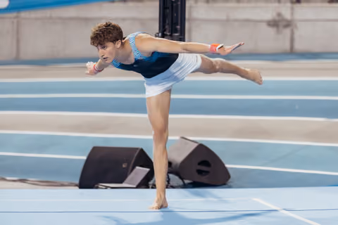 Young male gymnast holds a precise arabesque balance on a blue competition floor, arms extended with focused intensity.