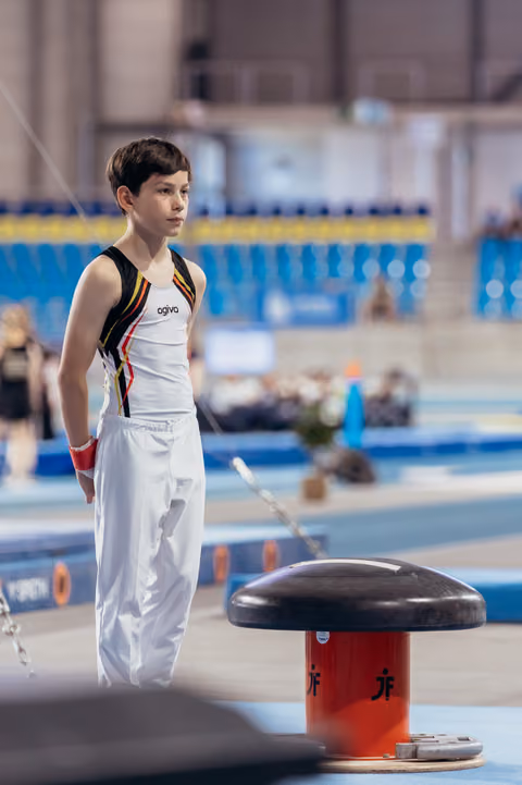 Young male gymnast stands focused and composed beside a pommel horse, wearing a Belgian-style leotard in a competition hall.