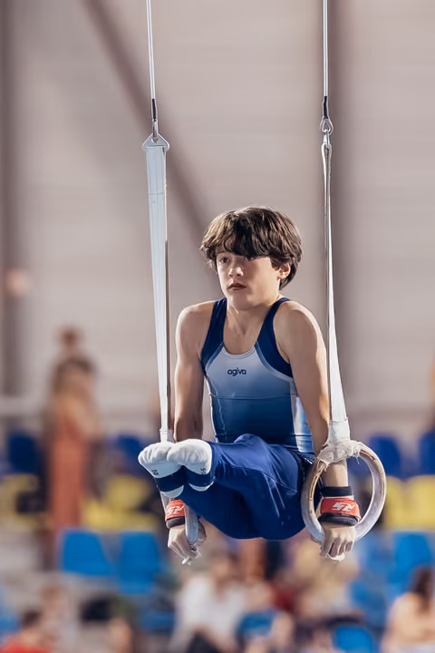 Young male gymnast performs an L-sit on still rings, expression focused and determined, in a competition gymnasium.