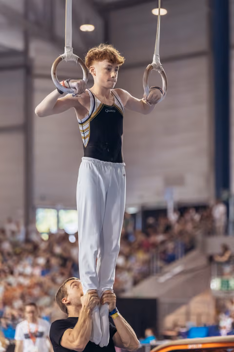 Focused young male gymnast grips still rings while a coach below steadies his feet, competing before a packed crowd.