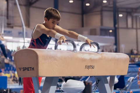 Focused young male gymnast grips pommel horse handles with concentration, wearing wrist guards at an indoor gymnastics meet.