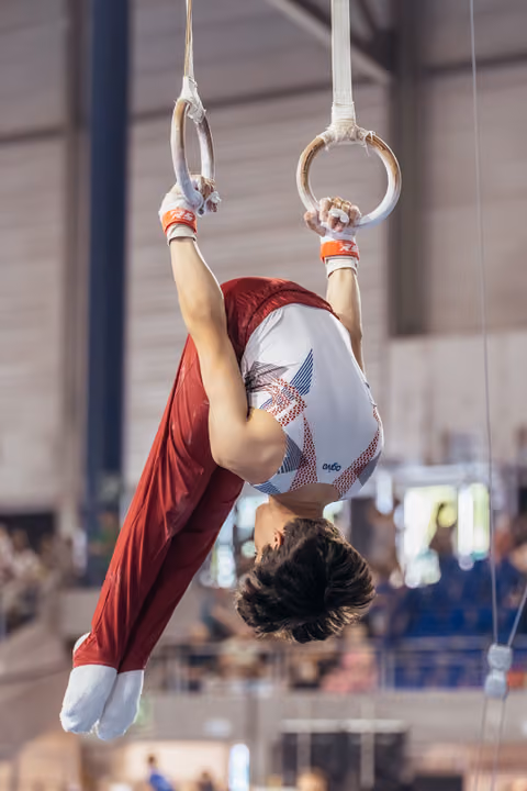 Young male gymnast performs an inverted hold on still rings, body curved with intense concentration in a competition hall.