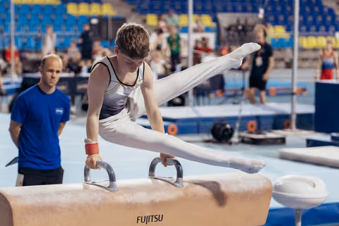 Young male gymnast performs a scissor split on the pommel horse, intense focus, while a coach watches nearby.