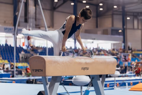 Young male gymnast performs on pommel horse, leg raised, intense focus during competition in packed indoor arena.