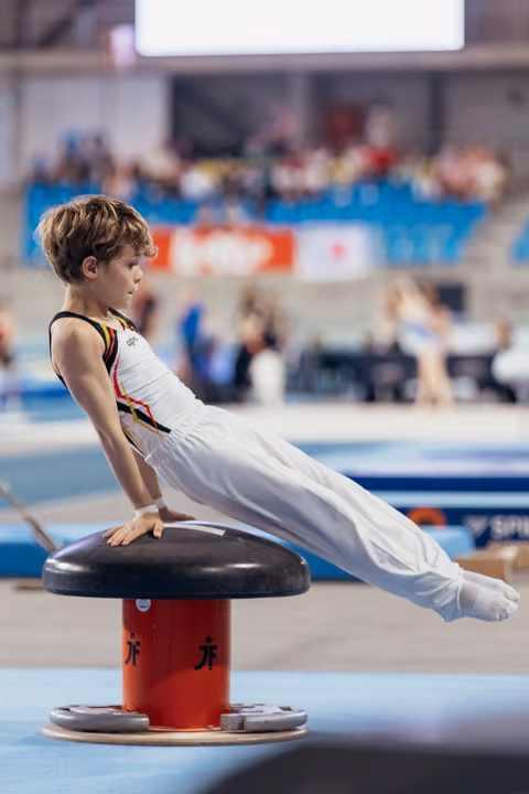 Young male gymnast balances on pommel mushroom with legs extended, showing intense focus during a gymnastics competition.