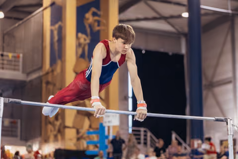 Young male gymnast grips parallel bars with fierce concentration, body horizontal, wearing red and blue leotard indoors.