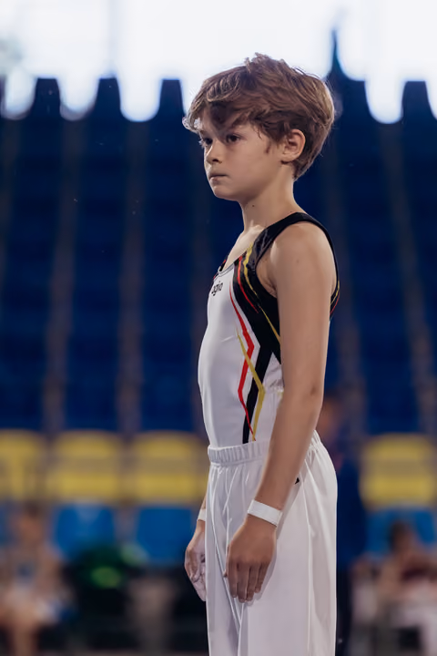 Young male gymnast stands still, focused expression, wearing white and black Belgian uniform in a competition arena.