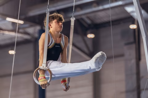 Young male gymnast holds steady on still rings, wearing Quatro leotard, legs raised, expression intense and focused.