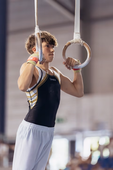 Young male gymnast grips still rings with intense focus, wearing a black Quatro leotard in a competition hall.