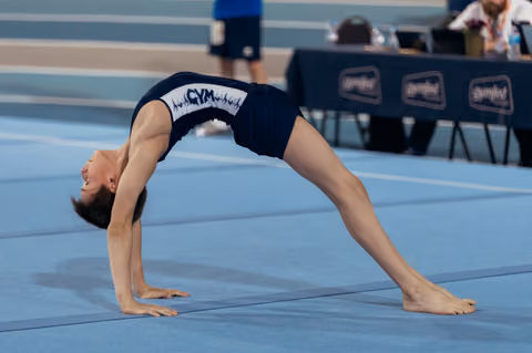 Young gymnast performs a backbend bridge on blue floor mat, head arched back, smiling during competition.