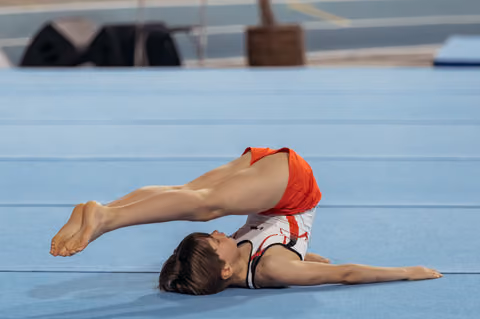 Gymnast performs a dramatic back arch on the floor exercise mat, legs raised high, face turned upward with focus.