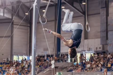 Young male gymnast hangs inverted on still rings, body arched with intense focus during a packed indoor gymnastics event.