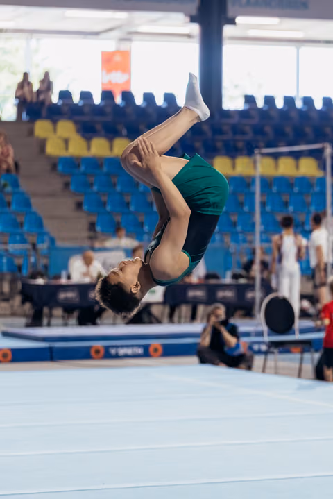 Young male gymnast executes a tight tucked backflip mid-air on the floor apparatus during an indoor gymnastics meet.
