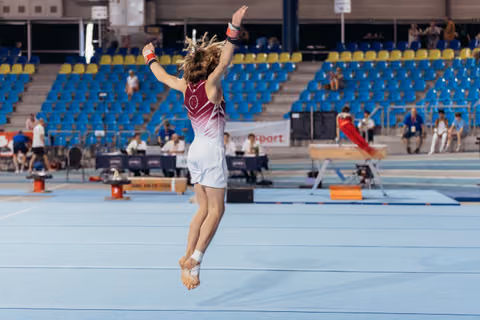 Young gymnast mid-jump on floor exercise, arms raised triumphantly, performing in a large indoor arena with blue seating.