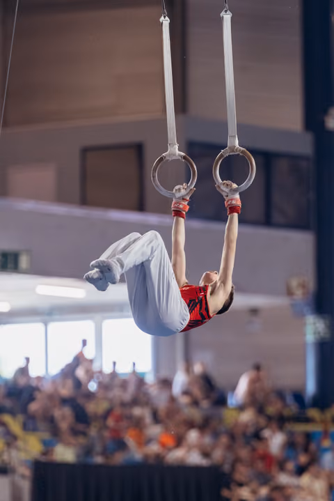 Young male gymnast performs an inverted hang on still rings, body arched, in front of a packed indoor arena crowd.