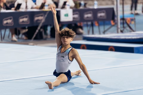 Young male gymnast holds a graceful seated pose on the floor mat, one arm raised high, focused expression.