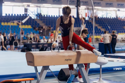 Young male gymnast performs on a Fujitsu pommel horse, focused and controlled, in a busy indoor gymnastics arena.