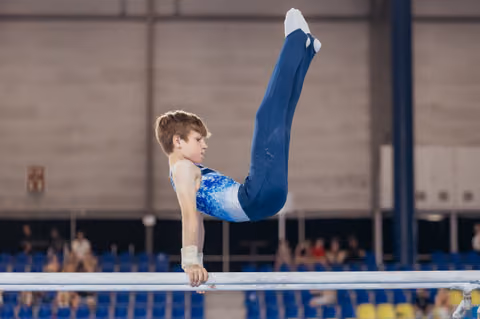 Focused young male gymnast performs an inverted hold on parallel bars, legs raised high, wearing a blue leotard.
