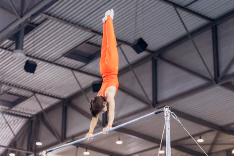 Male gymnast in orange performs a perfect handstand on the high bar, body fully extended, in an indoor gymnasium.