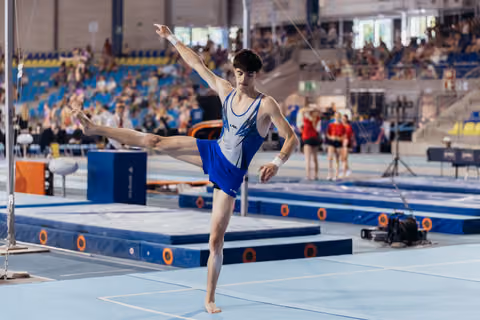 Young male gymnast performs a dynamic leg raise on the floor exercise, arms extended, focused expression in a busy arena.