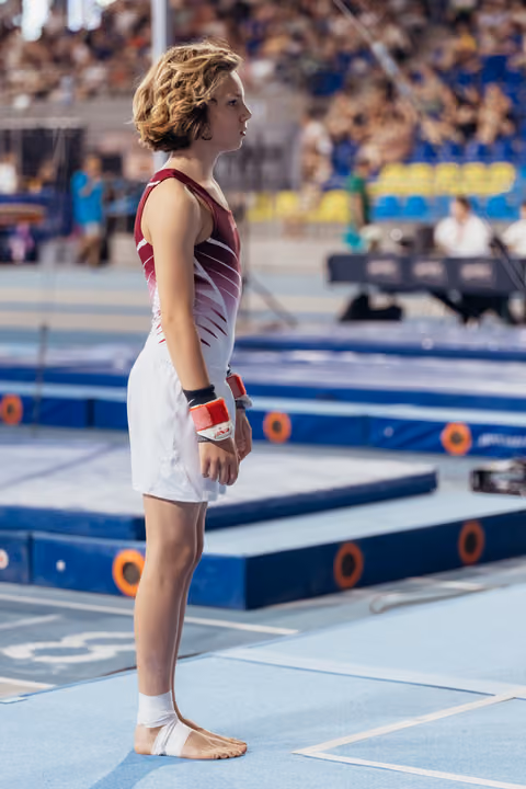 Young gymnast stands barefoot on blue floor mat, gazing forward with calm focus, wearing red leotard and white skirt.