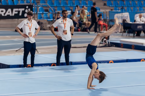 Young male gymnast performs a handstand on the floor while two coaches observe attentively in a blue competition arena.