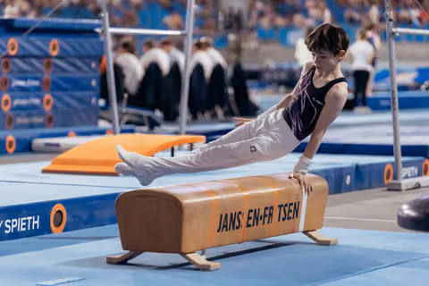 Young male gymnast performs a straddle hold on the pommel horse, legs extended, focused expression at an indoor competition.
