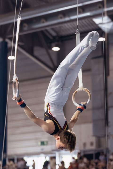 Young male gymnast performs an inverted pike hold on still rings, arms extended, body rigid with intense concentration.
