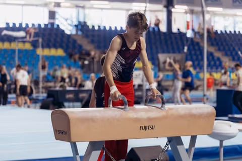 Young male gymnast grips pommel horse handles with focused concentration in a busy gymnastics arena