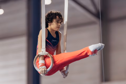 Young male gymnast holds still rings with intense focus, one leg raised, wearing red pants and navy leotard.
