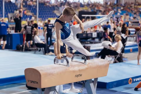 Young male gymnast performs on pommel horse, leg raised high, intense concentration, in a busy competition arena.