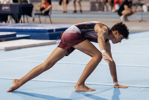 Young male gymnast barefoot on blue floor mat, crouched low in focused landing position during competition.