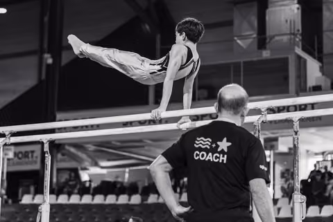 Young male gymnast performs a split hold on parallel bars while his coach observes attentively from below at a competition.