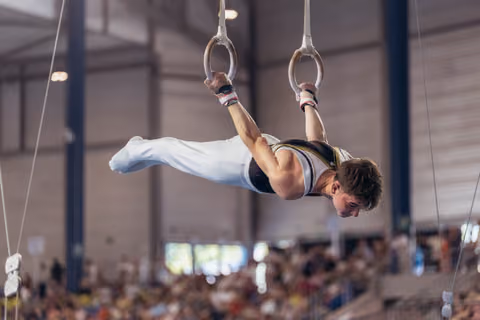 Young male gymnast performs an iron cross on still rings, body horizontal, intense focus, in a packed sports hall.