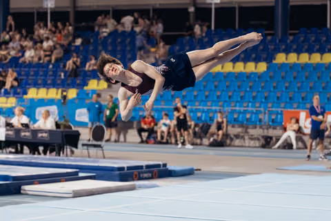 Young male gymnast soaring horizontally during floor exercise, body fully extended, in a packed indoor sports arena.