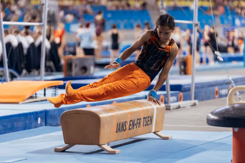 Young gymnast in orange uniform vaults over pommel horse with focused intensity at indoor gymnastics competition.