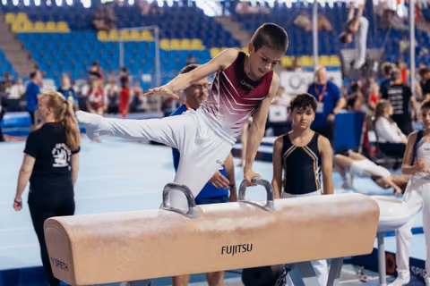 Young male gymnast performs on pommel horse, leaning forward with intense focus, competitors and coaches visible behind.
