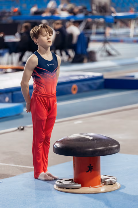 Young male gymnast in red and navy Quatro suit stands barefoot, eyes closed, composing himself before pommel horse routine.