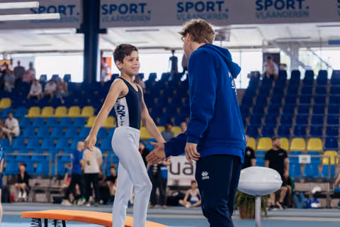 Smiling young gymnast shakes hands with coach in blue hoodie on the floor exercise mat in a packed arena.